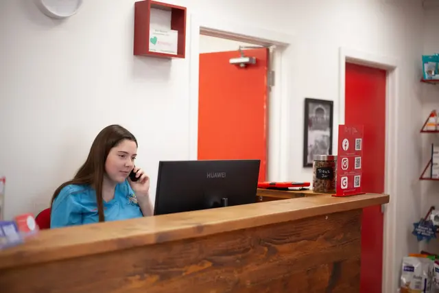 A young woman with long brown hair, wearing a light blue shirt, is sitting behind a wooden reception desk, talking on her cell phone and looking at a computer monitor.