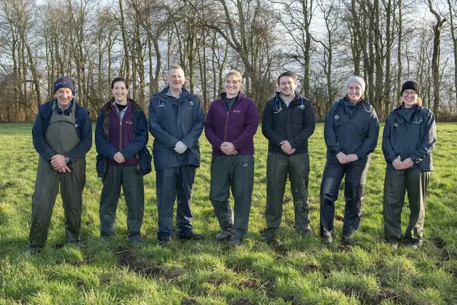 Group of seven people standing outdoors on a grassy field with trees in the background, dressed in outdoor and work clothes.