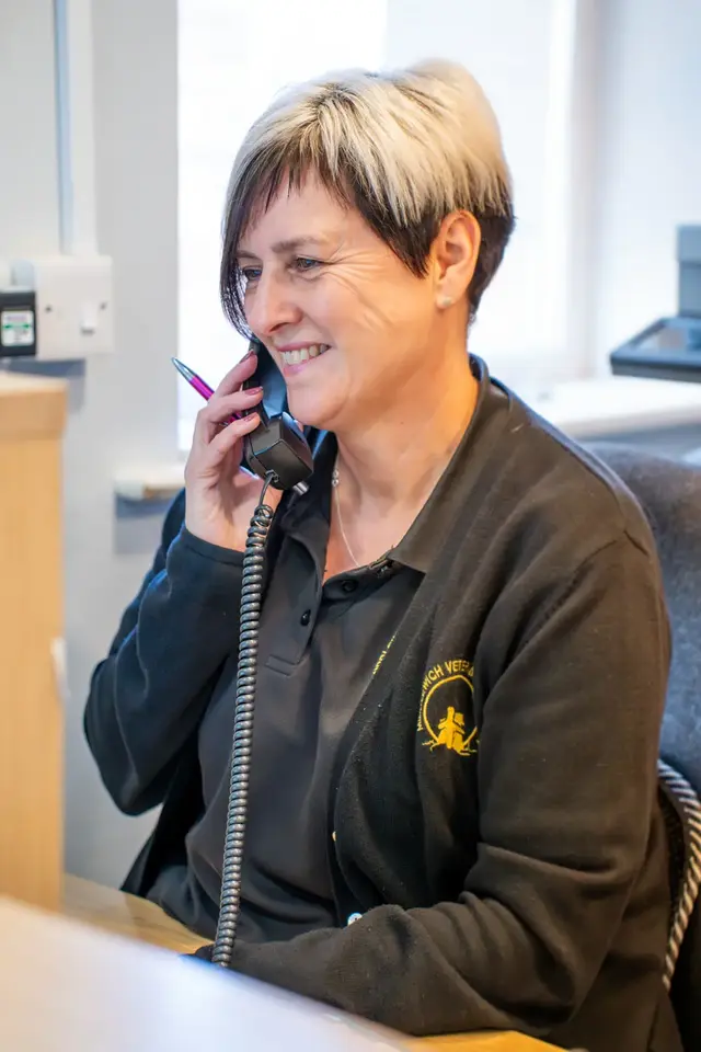 A woman with short hair is sitting at a desk in an office, smiling, and talking on a corded phone. She is wearing a black cardigan over a dark polo shirt.