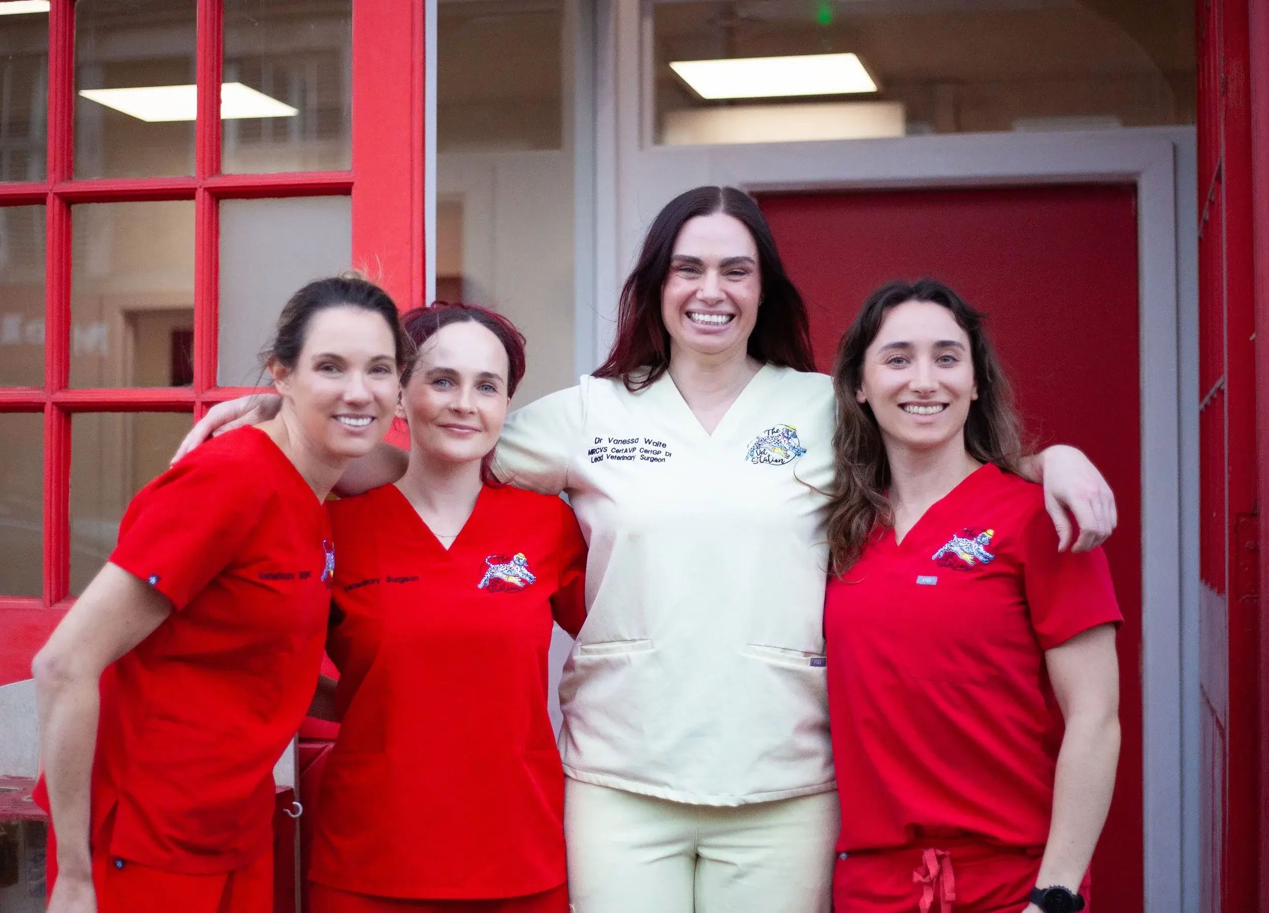 Four women, dressed in medical scrubs, stand together outside a building with red doors and windows, smiling for the photo.