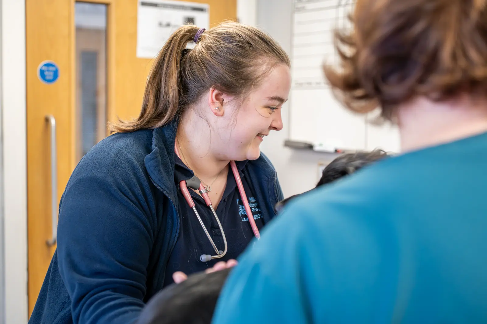A healthcare professional with a ponytail smiles while interacting with a person off-camera. She is wearing a stethoscope around her neck. The setting appears to be a medical office with a wooden door and a whiteboard in the background.