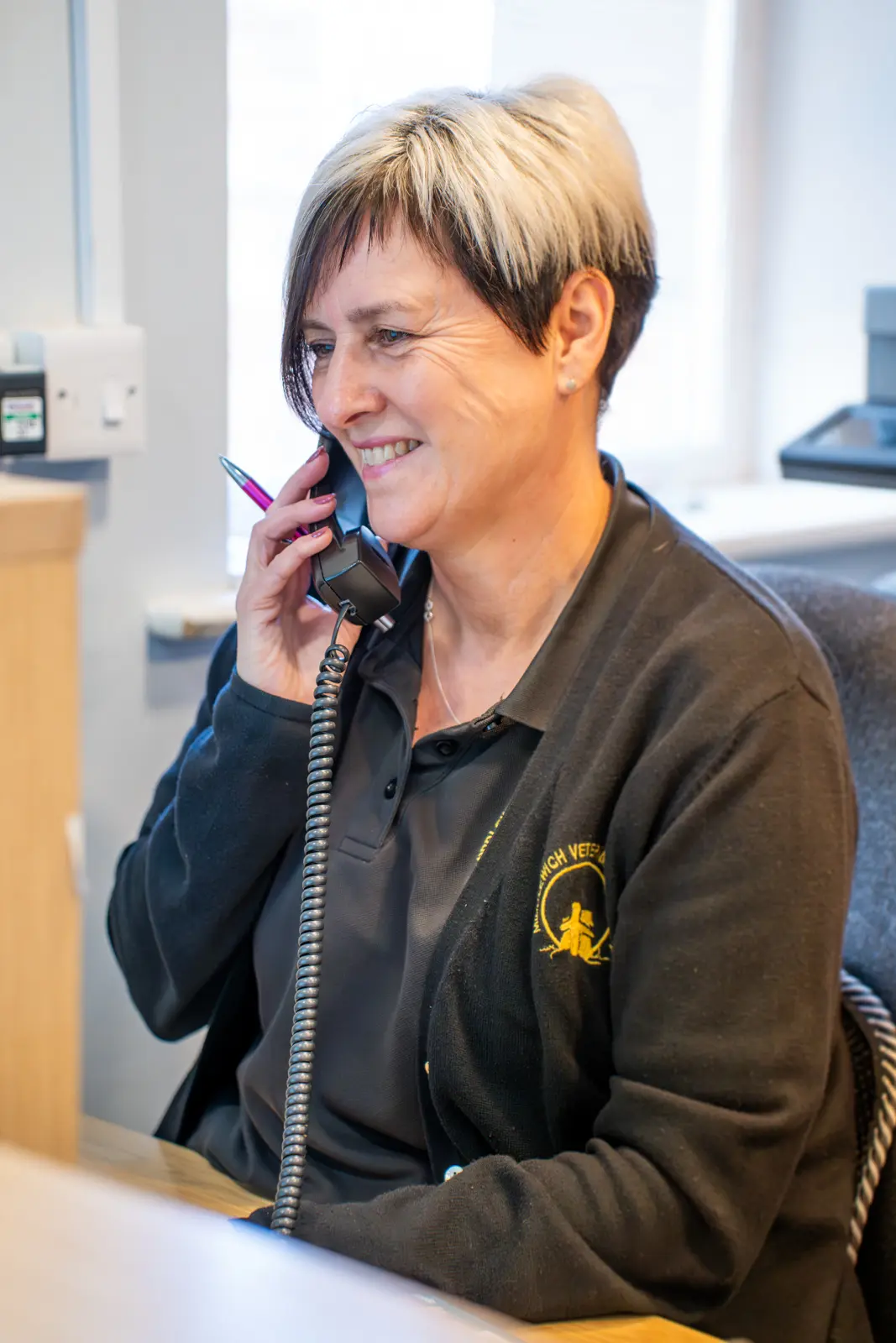 A woman with short hair is sitting at a desk in an office, smiling, and talking on a corded phone. She is wearing a black cardigan over a dark polo shirt.